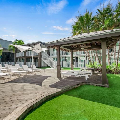 An outdoor area with lounge chairs, a wooden deck, shaded seating area, artificial grass, and tropical plants, adjacent to two-story building.