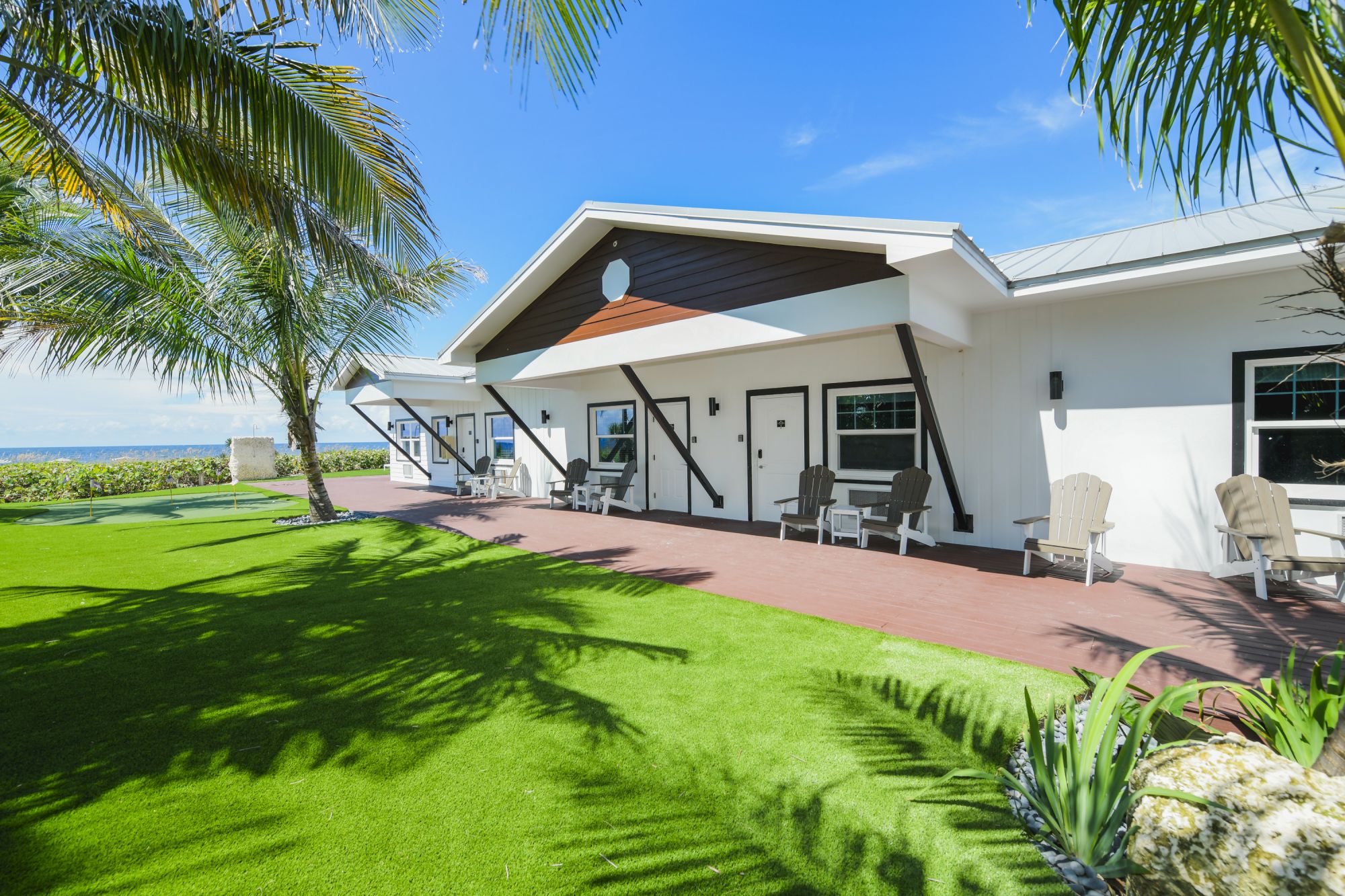 A modern bungalow with a lush green lawn, palm trees, and a patio with chairs, under a clear blue sky.