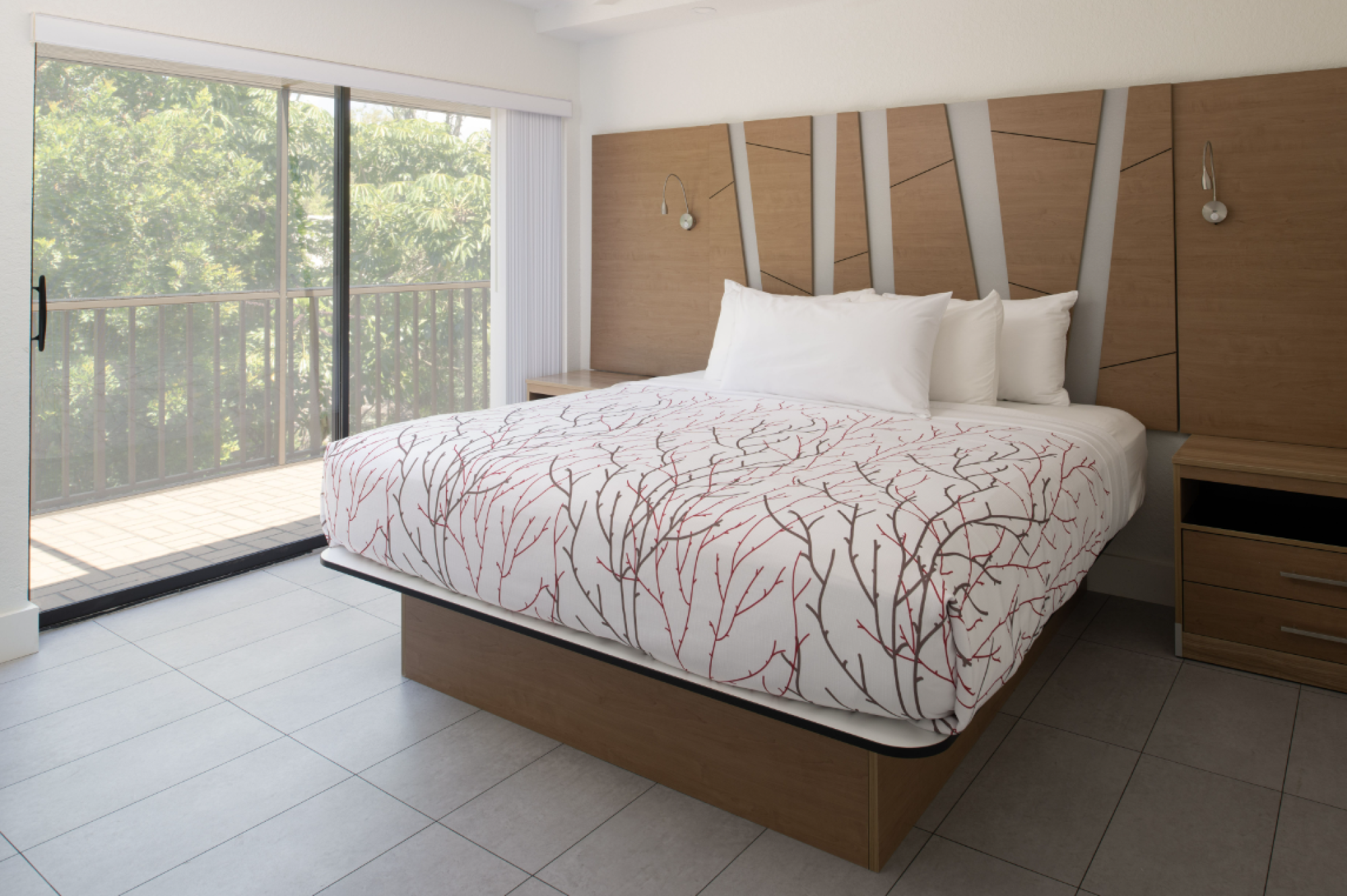 A modern bedroom features a large bed with white and red patterned bedding, nightstands, and a sliding glass door leading to a balcony with a railing.