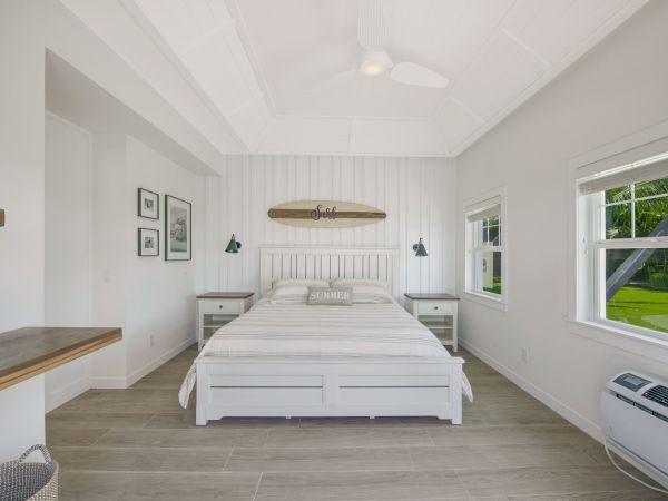 A bright bedroom with a white bed, surfboard wall decor, two nightstands, open windows, and a ceiling fan. There's also a wicker basket and an AC unit.