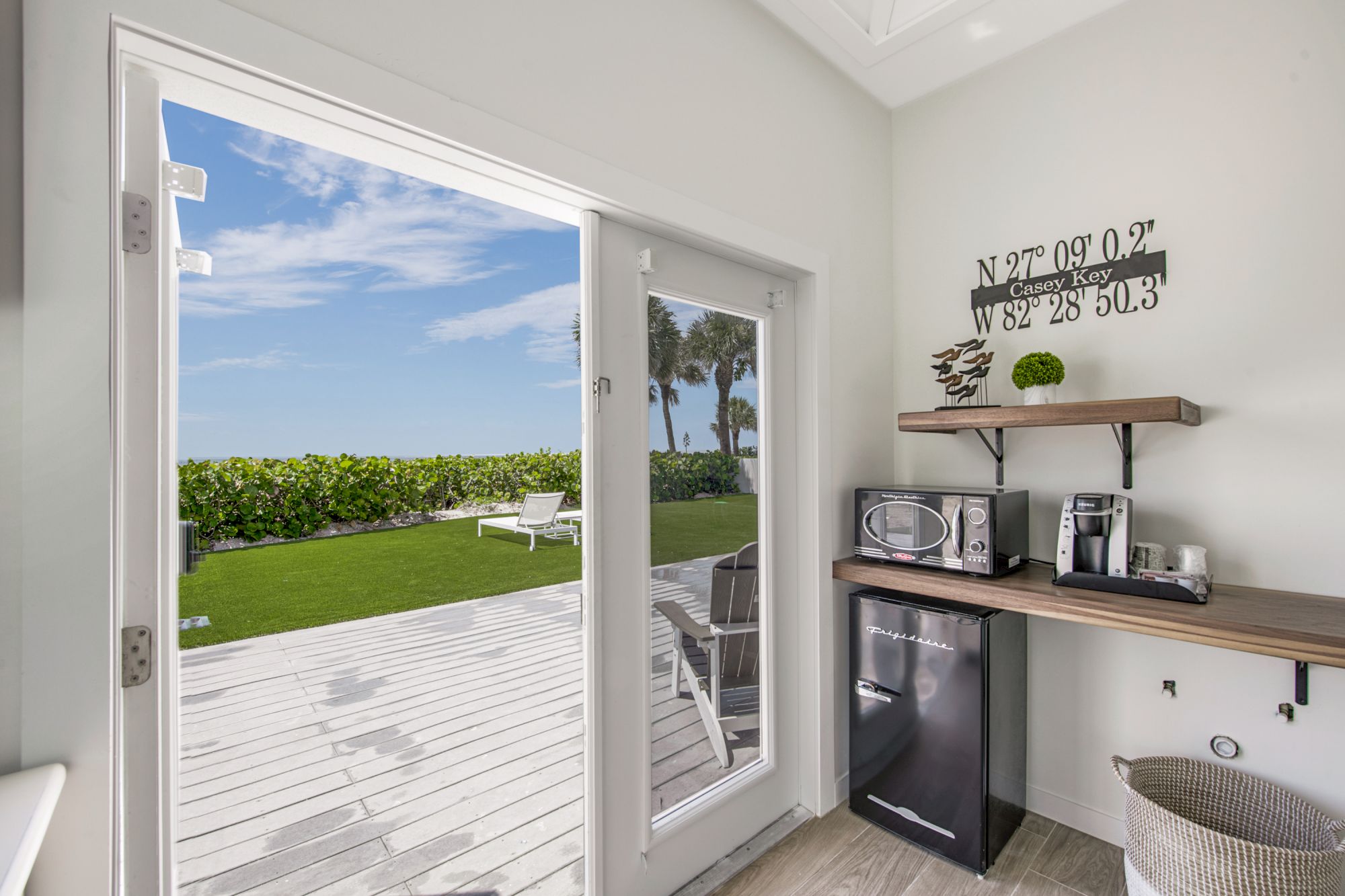 A small kitchenette with a refrigerator, microwave, and coffee maker opens to a deck with a lawn and ocean view through a sliding glass door.