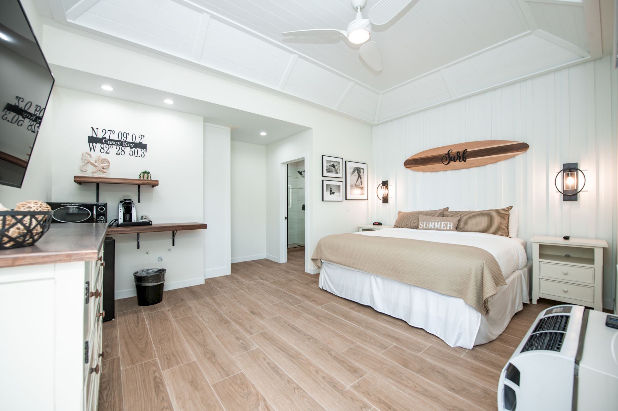 A cozy bedroom with a modern coastal theme, featuring a large bed, wall-mounted TV, surfboard wall decor, and light wood floors ending the sentence.