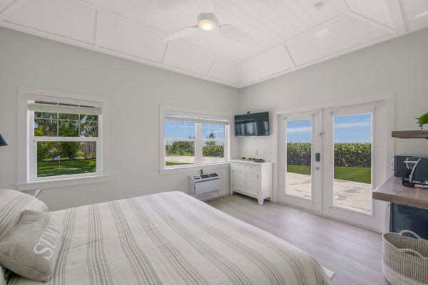 A bright bedroom with a striped bedspread, windows, a TV, desk, and glass doors opening to a patio with an ocean view.