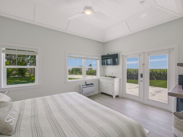 A bright bedroom with a striped bedspread, windows, a TV, desk, and glass doors opening to a patio with an ocean view.