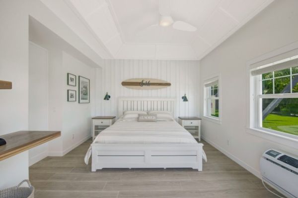 A bright bedroom with a white bed, surfboard decor, side tables, green lamps, window, wooden floor, and air conditioning unit ends the sentence.