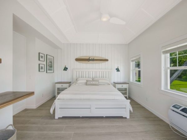 A bright bedroom with a white bed, surfboard decor, side tables, green lamps, window, wooden floor, and air conditioning unit ends the sentence.