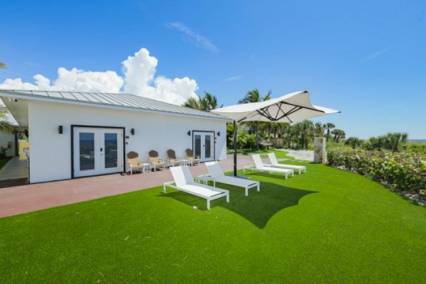 A modern white house with lounge chairs on a green lawn and a large umbrella on a sunny day.