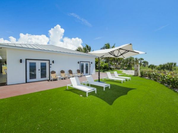 A modern white house with lounge chairs on a green lawn and a large umbrella on a sunny day.