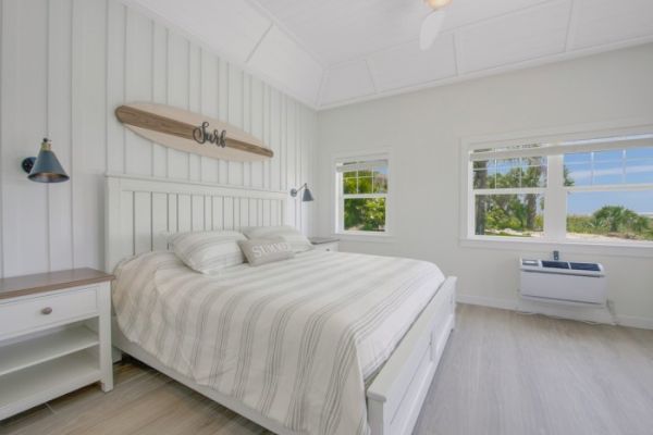 A bright bedroom with white decor, a large bed, two windows showing a greenery view, two bedside tables, wall lamps, and an air conditioner.