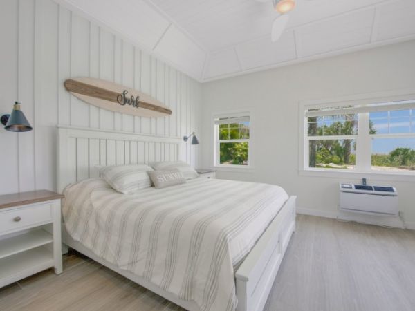 A bright bedroom with white decor, a large bed, two windows showing a greenery view, two bedside tables, wall lamps, and an air conditioner.