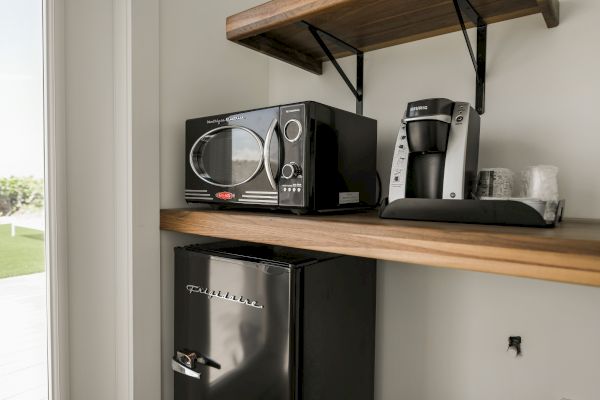 The image shows a kitchen countertop with a microwave, coffee maker, mini fridge, and cups on a wooden shelf, next to a window.