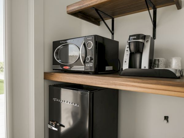 The image shows a kitchen countertop with a microwave, coffee maker, mini fridge, and cups on a wooden shelf, next to a window.