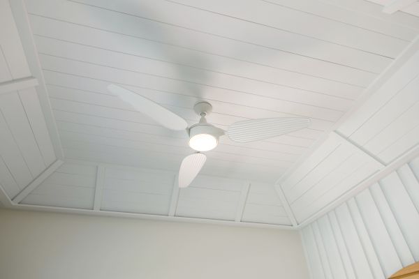 The image shows a white ceiling with a white ceiling fan that has a central light, under a paneled roof.