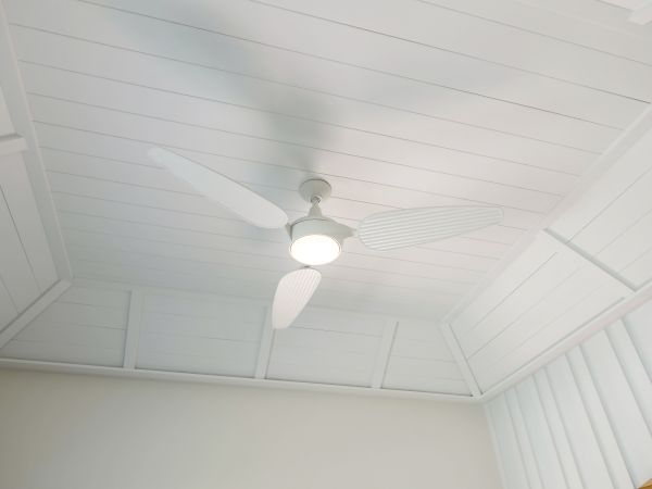 The image shows a white ceiling with a white ceiling fan that has a central light, under a paneled roof.