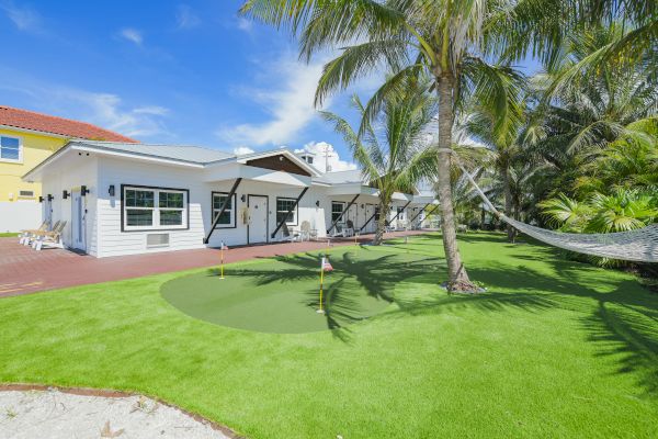 A white house with a red roof sits beside a well-maintained lawn featuring a small putting green, palm trees, and a hammock.