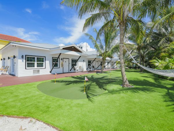 A white house with a red roof sits beside a well-maintained lawn featuring a small putting green, palm trees, and a hammock.