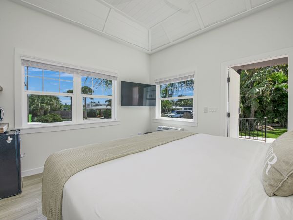 A bright bedroom with white walls, a bed, TV, window views of a garden, a mini-fridge, and a door opening to the outside is shown.