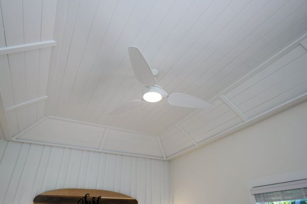 A white ceiling fan with light is mounted on a white-paneled, vaulted ceiling in a room with white walls and a window, ending the sentence.