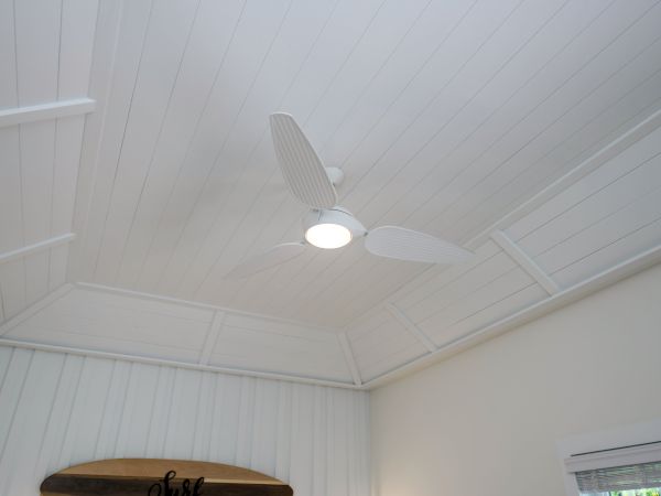 A white ceiling fan with light is mounted on a white-paneled, vaulted ceiling in a room with white walls and a window, ending the sentence.