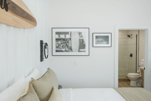 A bedroom with a bed, striped pillows, wall-mounted surfboard, framed pictures, an open door leading to a bathroom with a toilet and towels.