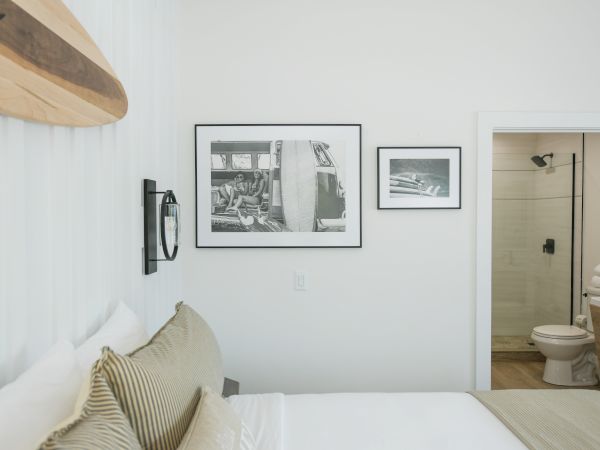 A bedroom with a bed, striped pillows, wall-mounted surfboard, framed pictures, an open door leading to a bathroom with a toilet and towels.