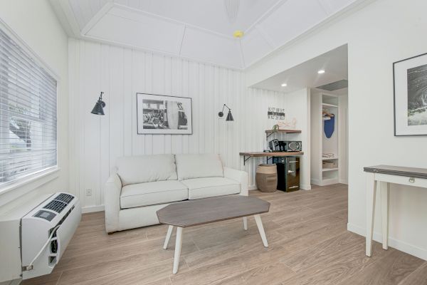 A modern, minimalistic living space with a sofa, coffee table, wall-mounted lights, framed pictures, and a small kitchenette area in the background.