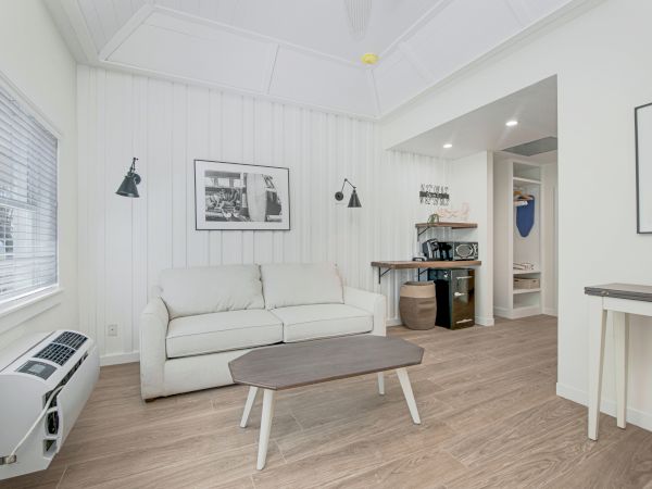 A modern, minimalistic living space with a sofa, coffee table, wall-mounted lights, framed pictures, and a small kitchenette area in the background.