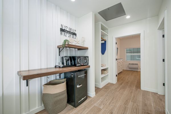 A modern room features a small fridge, microwave, decorative sign, basket, open shelving, and wood flooring, leading into an adjacent bedroom.