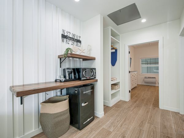A modern room features a small fridge, microwave, decorative sign, basket, open shelving, and wood flooring, leading into an adjacent bedroom.