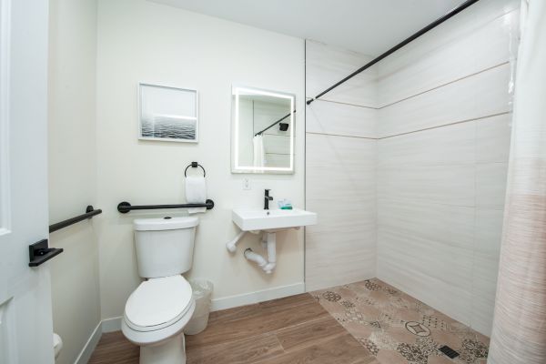 A modern bathroom with a toilet, sink, mirror, towel holder, and a walk-in shower, featuring wood flooring and minimalistic decor.