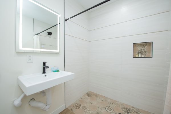 This image shows a minimalist bathroom with a white sink, a mirror, and a spacious walk-in shower with a patterned floor and a small niche.