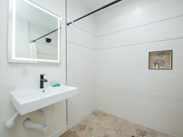 This image shows a minimalist bathroom with a white sink, a mirror, and a spacious walk-in shower with a patterned floor and a small niche.