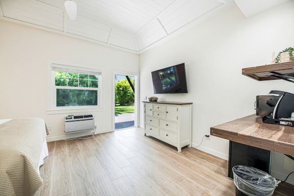 A bright, clean room featuring a wall-mounted TV, bed, dresser, air conditioning unit, desk with coffee maker, and a door leading outside.