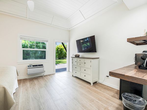 A bright, clean room featuring a wall-mounted TV, bed, dresser, air conditioning unit, desk with coffee maker, and a door leading outside.