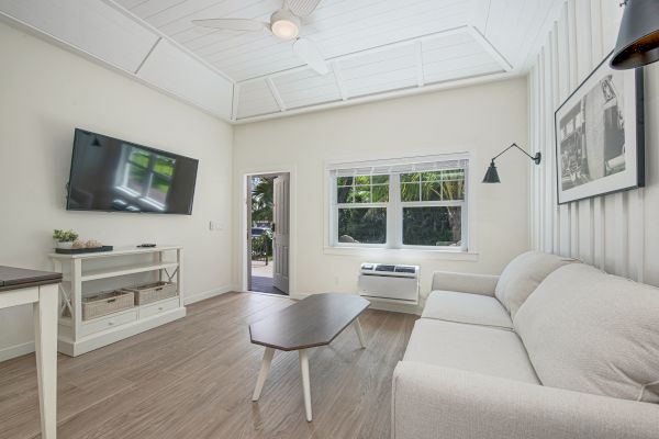 A modern living room with a wall-mounted TV, a beige sofa, a wooden coffee table, and a large window bringing in natural light.