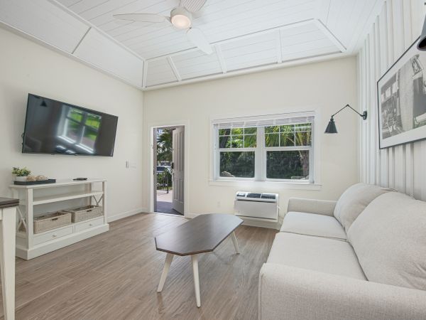 A modern living room with a wall-mounted TV, a beige sofa, a wooden coffee table, and a large window bringing in natural light.