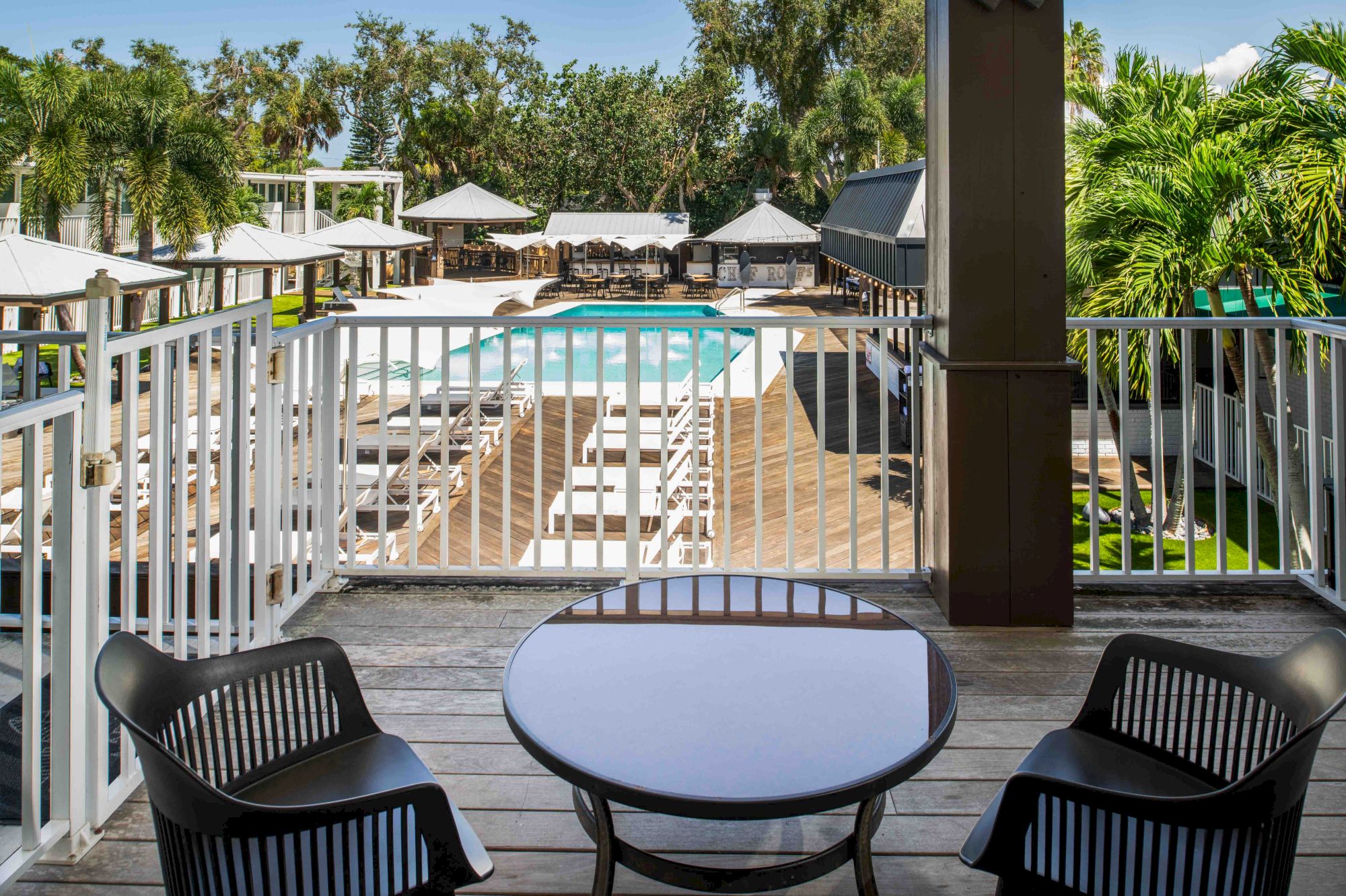 A view of a pool area with lounge chairs and umbrellas, taken from a balcony with a table and two chairs, surrounded by palm trees and greenery.