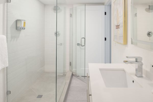 The image shows a modern, white bathroom with a glass-enclosed shower, a sleek vanity with a rectangular sink, a mirror, and a wall-mounted faucet.