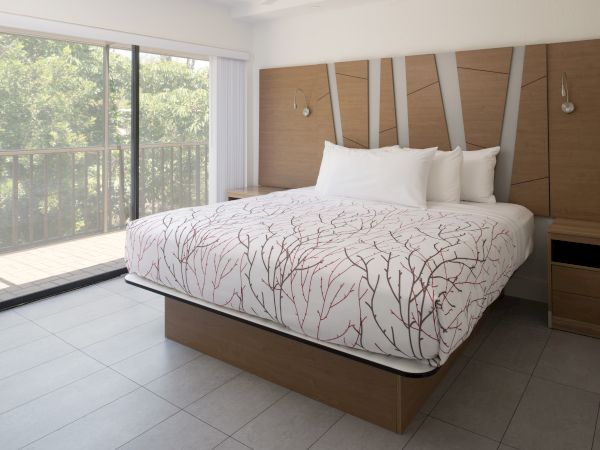 A modern bedroom with a platform bed, white bedding with a branch pattern, wooden headboard, bedside tables, and a sliding glass door leading to a balcony.