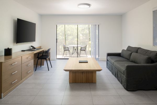 The image shows a modern living room with a sofa, coffee table, TV, work desk, and sliding glass doors opening to an outdoor area.