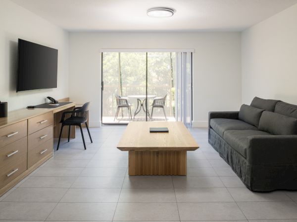 The image shows a modern living room with a sofa, coffee table, TV, work desk, and sliding glass doors opening to an outdoor area.