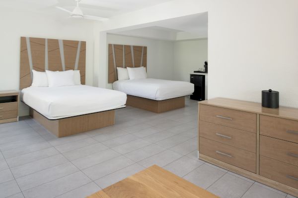 The image shows a minimalistic hotel room with two neatly made beds, a wooden dresser, and tiled flooring, featuring a clean and modern design.