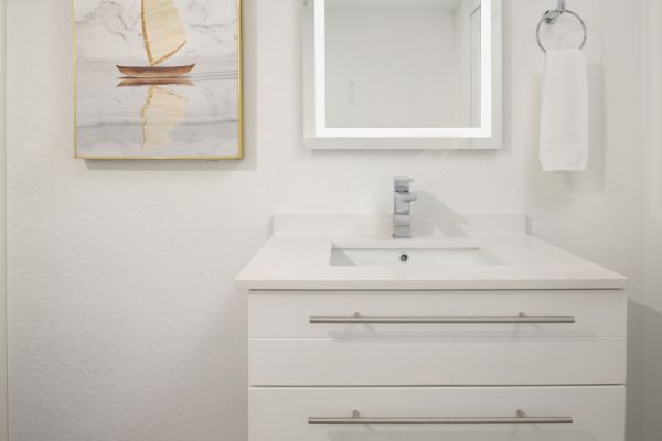 A modern bathroom with a white vanity, a square mirror, a towel ring with a white towel, and a painting of a sailboat on the wall.