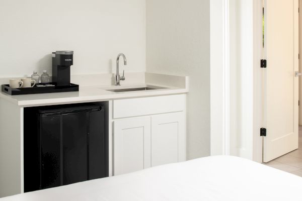 A small kitchenette with a sink, cabinets, a black mini-fridge, and a coffee maker on the counter beside a tray with cups and condiments.