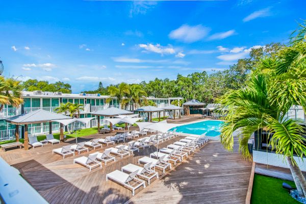 A luxury resort pool area with lounge chairs, umbrellas, palm trees, and modern buildings set against a blue sky and lush greenery.