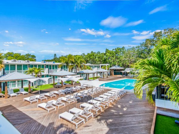 A luxury resort pool area with lounge chairs, umbrellas, palm trees, and modern buildings set against a blue sky and lush greenery.