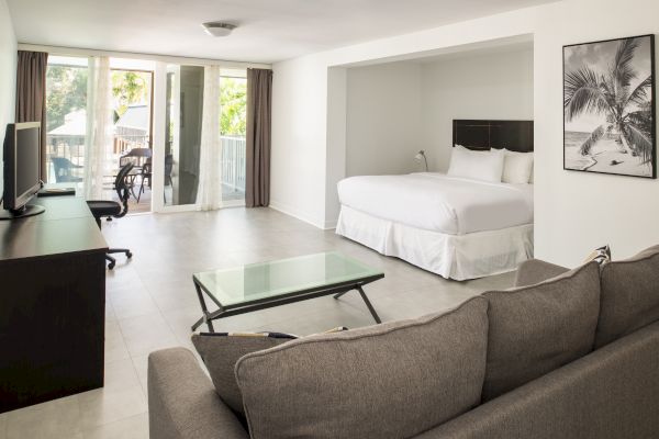 A spacious hotel room with a bed, sofa, TV, desk, and glass coffee table. There's a balcony with curtains, and a framed wall art piece.