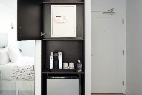 The image shows a hotel room with a closet containing a safe, a coffee maker, cups, a bottle of water, and a mini fridge, next to a closed door.