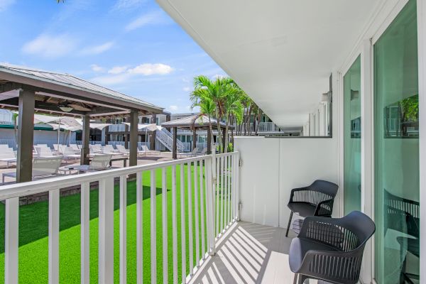 A balcony with two chairs overlooks a pool area with lounge chairs and umbrellas, surrounded by lush greenery and clear blue skies.