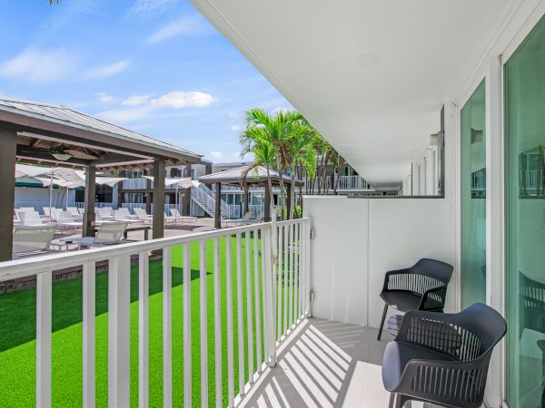 A balcony with two chairs overlooks a pool area with lounge chairs and umbrellas, surrounded by lush greenery and clear blue skies.